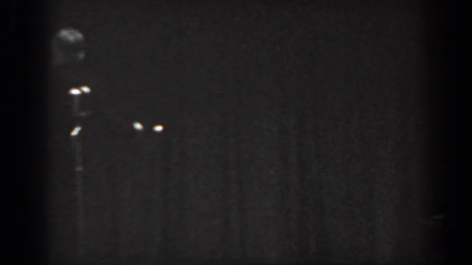 Black-and-white 8mm home movie frame from a 1965 stage performance, showing a distant figure and two stage-light glints against a near-black curtain backdrop.