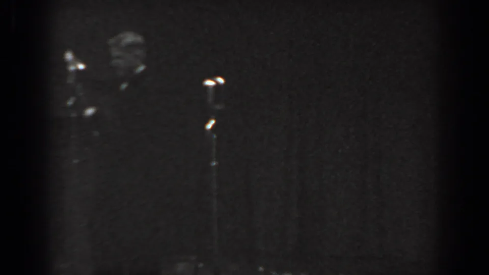 Black-and-white 8mm home movie frame, circa 1965, showing two shadowy figures and a clearly visible floor-standing microphone stand on a dimly lit talent-show stage.