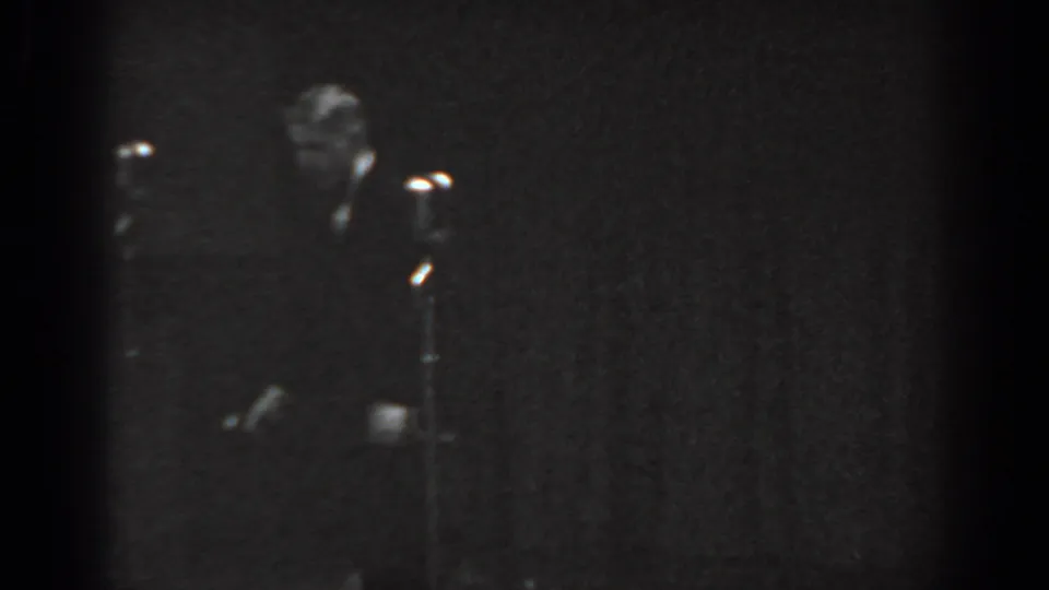 Black-and-white 8mm home movie frame from a 1965 talent show, showing a figure in dark attire at stage right and a floor-standing microphone stand against a dark curtain backdrop.