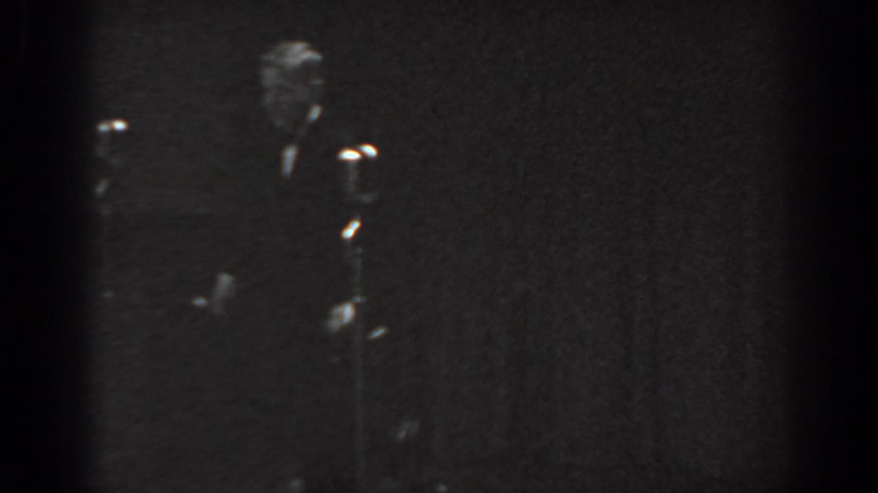 Black-and-white 8mm frame from a 1965 Saint Louis stage performance; a formally dressed man stands near a vintage microphone stand under dim stage lighting.