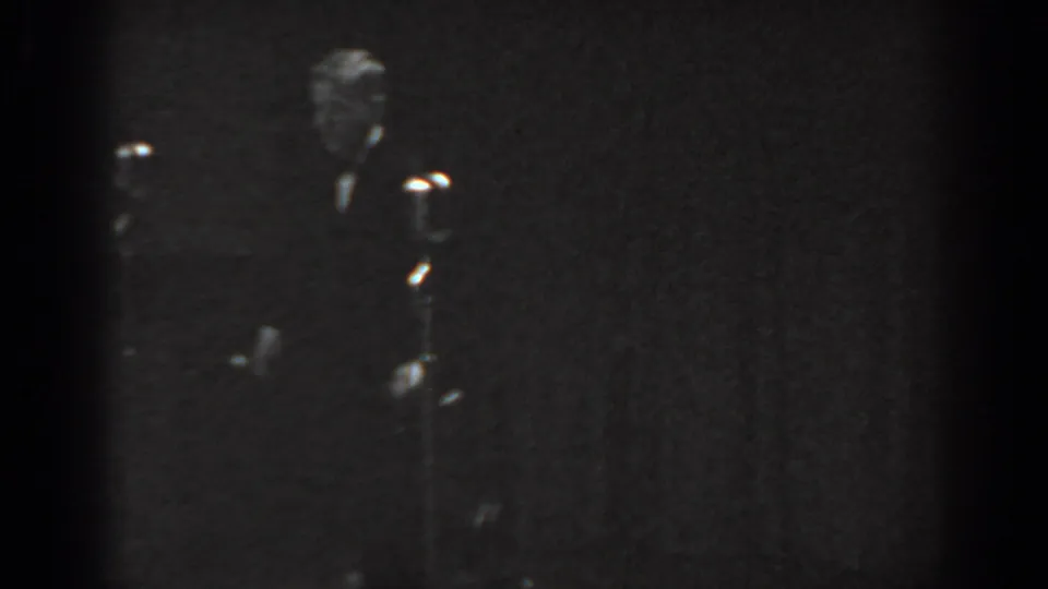 Black-and-white 8mm frame from a 1965 Saint Louis stage performance; a formally dressed man stands near a vintage microphone stand under dim stage lighting.