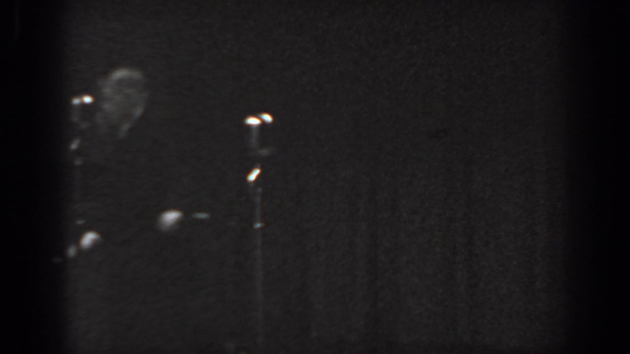 Formally dressed man visible at upper left of a dark stage with a microphone stand to the right under low-key lighting.