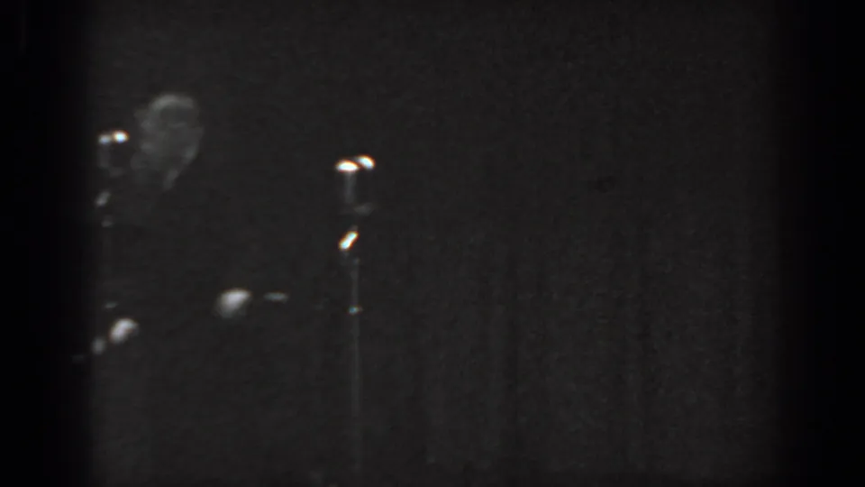 Black-and-white 8mm frame from an apparent 1965 stage performance; formally dressed performer near a vintage microphone stand against a uniformly dark backdrop.
