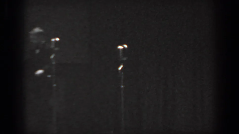 Black-and-white 8mm frame from a 1965 Saint Louis stage act; performer at lower-left with a vintage microphone stand and illuminated capsule tip in the foreground.