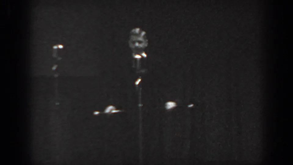 Black-and-white 8mm frame from a 1965 stage performance; two vintage microphone stands visible with a performer largely obscured by heavy underexposure.