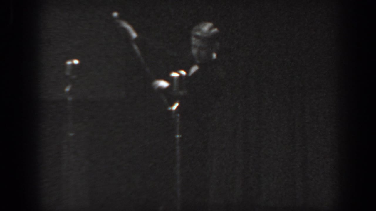 8mm home movie frame circa 1965 showing a man in dark suit raising a cane on stage, with a microphone stand at left and dark curtain behind.