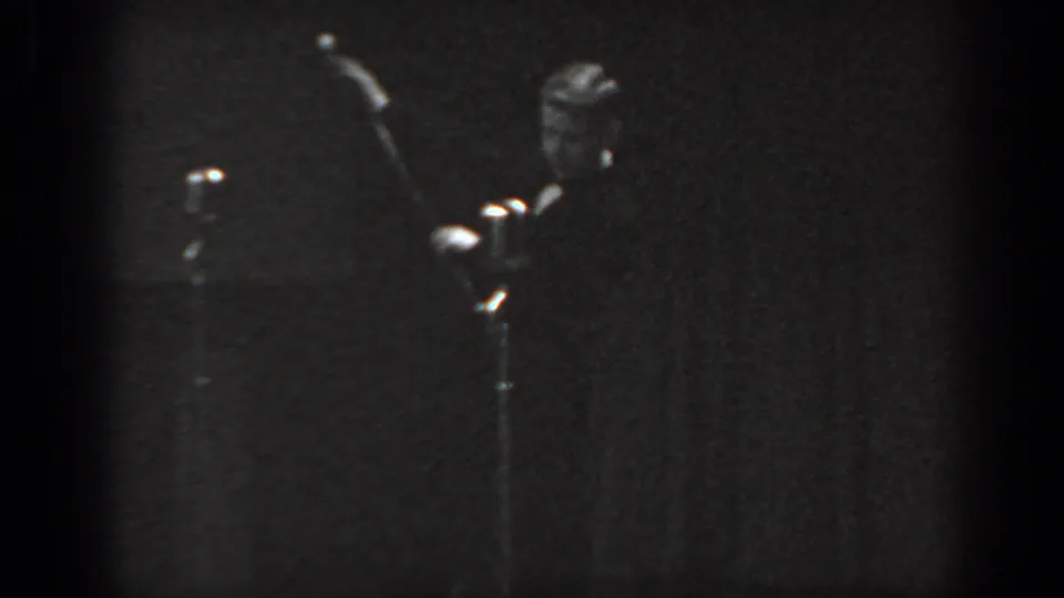 8mm home movie frame circa 1965 showing a man in dark suit raising a cane on stage, with a microphone stand at left and dark curtain behind.