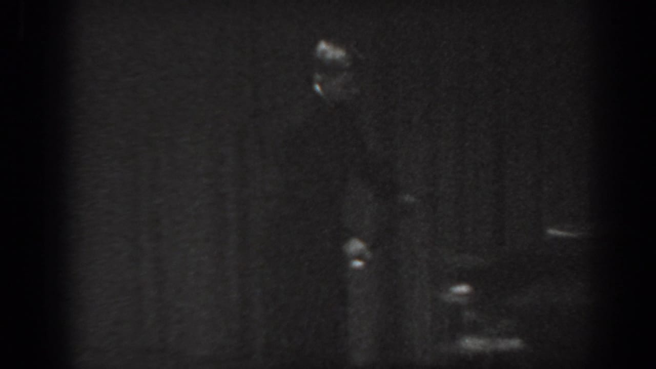 Man in dark suit standing on stage against a dark curtain backdrop; left portion of frame heavily underexposed.
