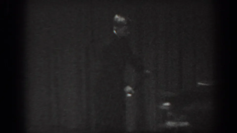 8mm home movie frame circa 1965 showing a man in dark suit standing on a dimly lit stage with a dark curtain backdrop.