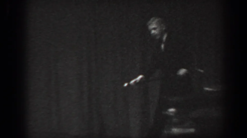 8mm home movie frame circa 1965 showing a man in dark suit bent into a dance pose on stage, cane prop visible, chairs partially seen below.