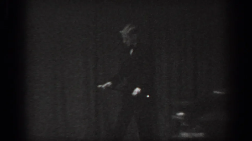 8mm home movie frame circa 1965 showing a man in dark suit in motion at right of frame, chairs and dark curtain visible in background.