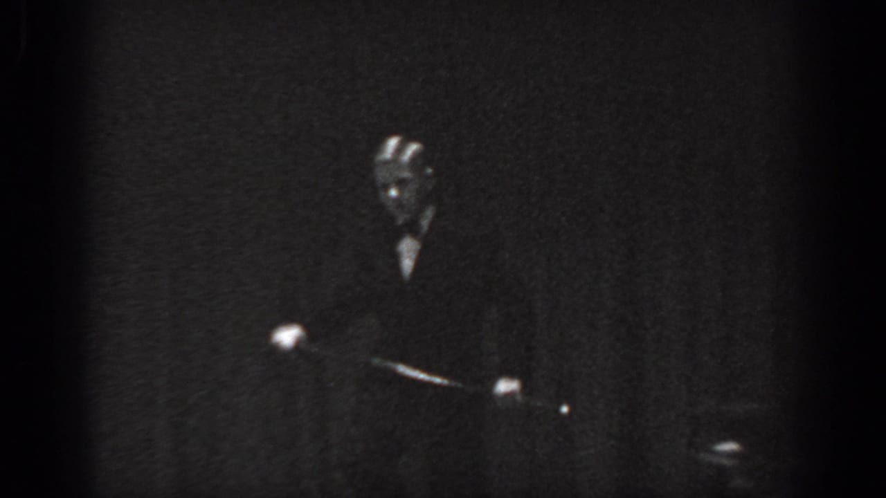 Male performer standing upright in dark formal attire on a dimly lit stage, spotlight visible, 8mm black-and-white.