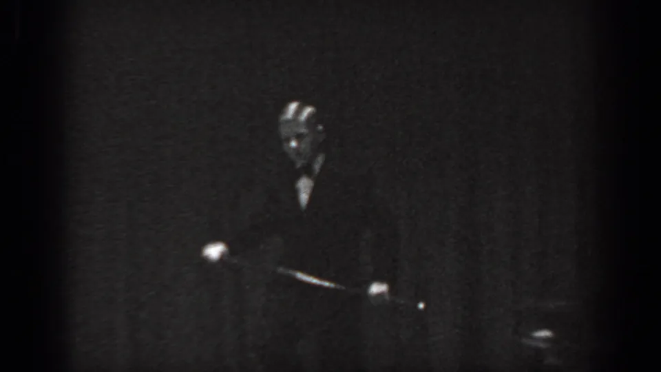 Cabaret performer standing center-stage in dark suit under spot lighting, appears to extend an arm or prop, Saint Louis, Missouri, 1965, 8mm film.