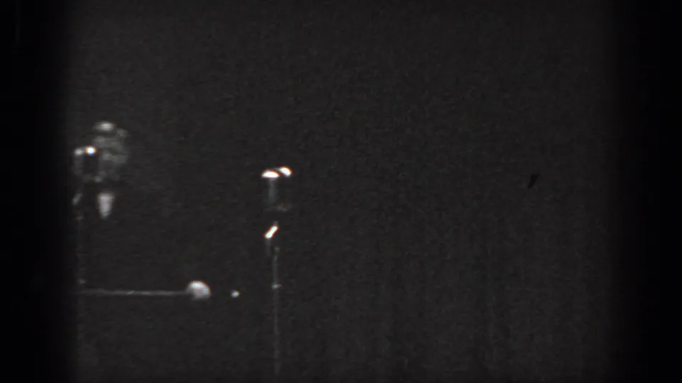 Cabaret performer standing on dark stage, possible microphone stand visible at right with point light sources below, Saint Louis, Missouri, 1965, 8mm film.