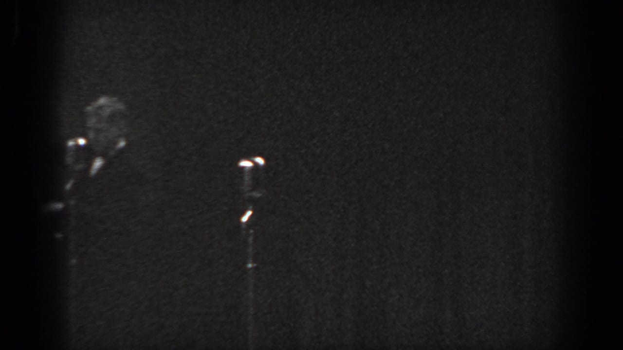 Grainy 8mm home-movie frame circa 1965, showing a man in a dark suit partially visible at stage left beside a stand microphone with a bright head.