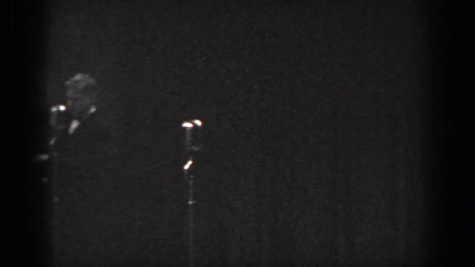 Grainy 8mm home-movie frame circa 1965, showing a man in a dark suit partially visible at stage left beside a stand microphone with a bright head.