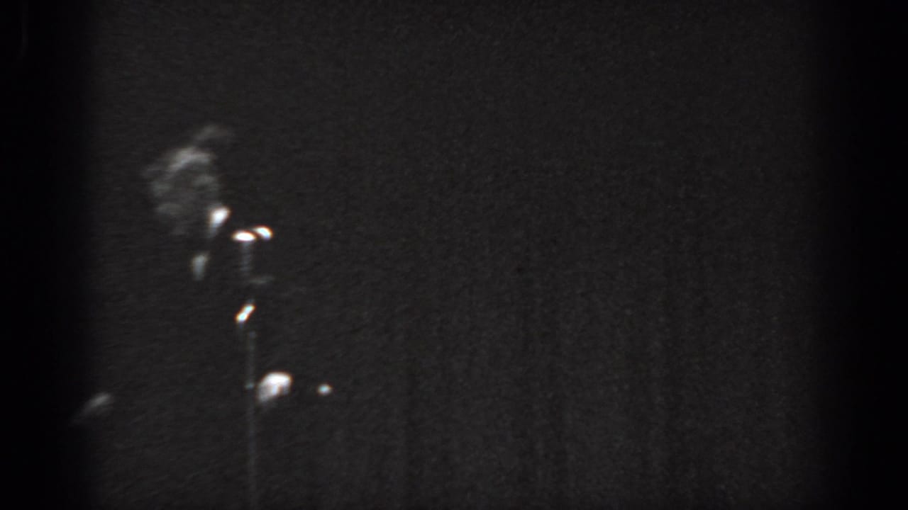 Dark 8mm frame with a figure in a suit more visible at upper left, a lit stand microphone, and a secondary point of stage light visible nearby.