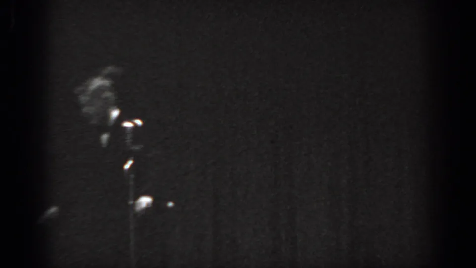 Grainy 8mm frame circa 1965, Saint Louis, showing a performer in a dark suit near a stand microphone with stage backlighting creating a partial silhouette.