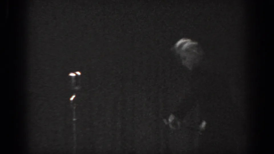 Grainy 8mm frame circa 1965, Saint Louis, with a performer visible at frame right near a lit stand microphone on a dark stage.