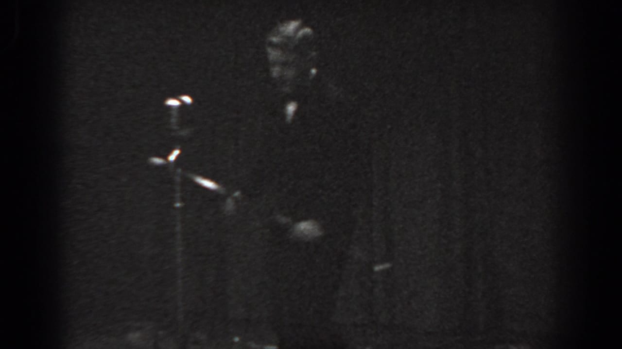 Young child in dark formal suit standing at a vintage microphone stand on a dimly lit stage, black-and-white 8mm film.