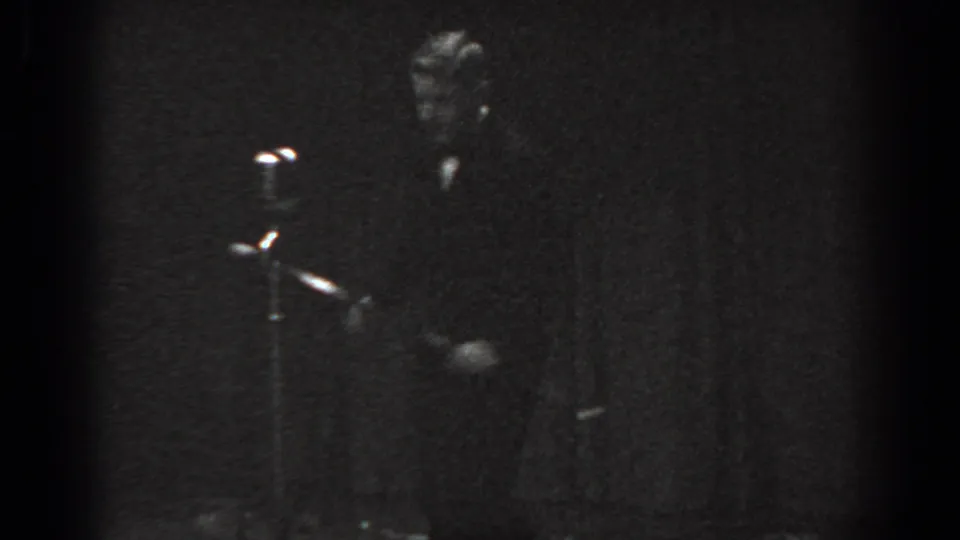 Child performer in dark suit beside a floor-standing vintage microphone stand on a low-lit stage, circa 1965 8mm home movie, Saint Louis Missouri.