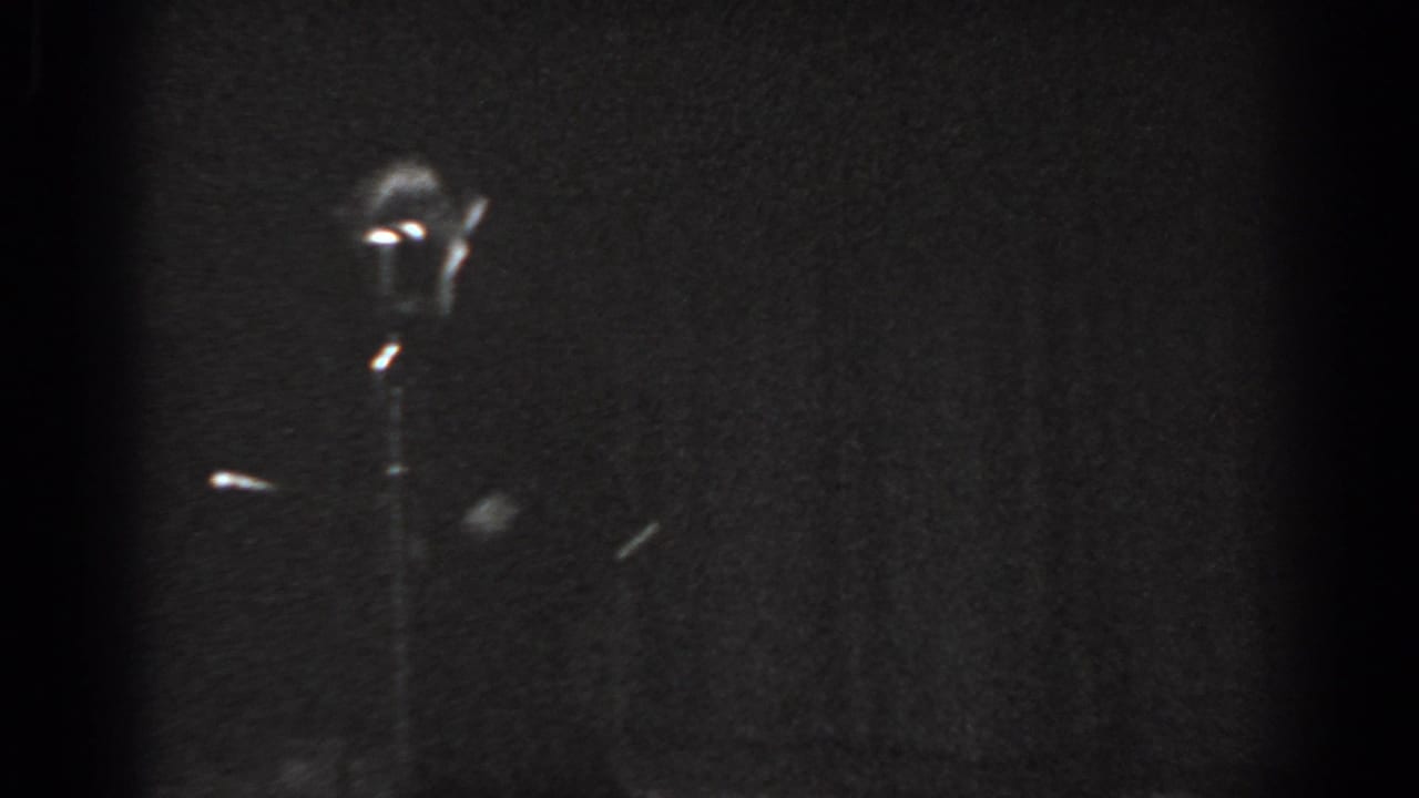 Child in motion beside a microphone stand on a dark stage, motion blur on limbs, grainy black-and-white 8mm film.