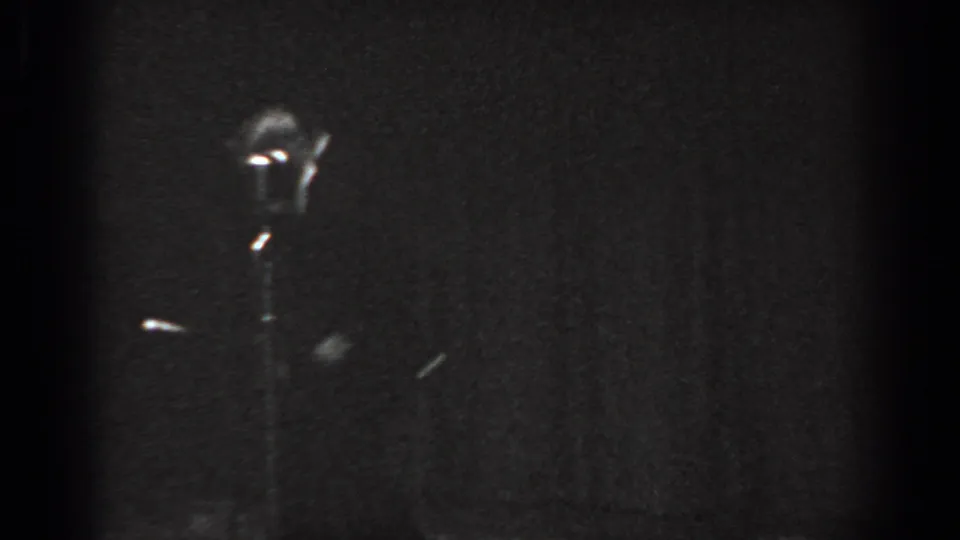 Child tap dancer mid-movement beside a vintage microphone stand on a dark stage, circa 1965 8mm home movie, Saint Louis Missouri.