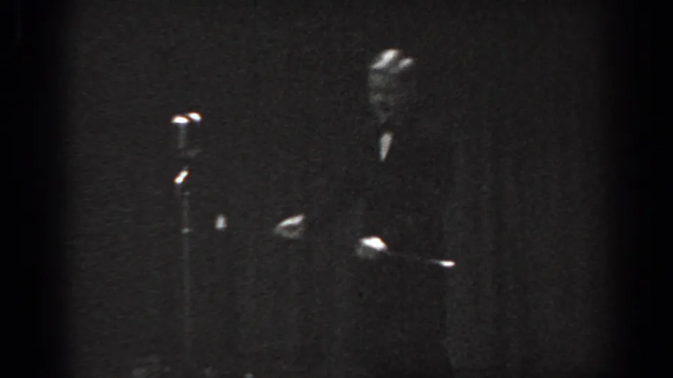 Child tap dancer in a low dance move on a dimly lit stage with vintage microphone stand, circa 1965 8mm home movie.