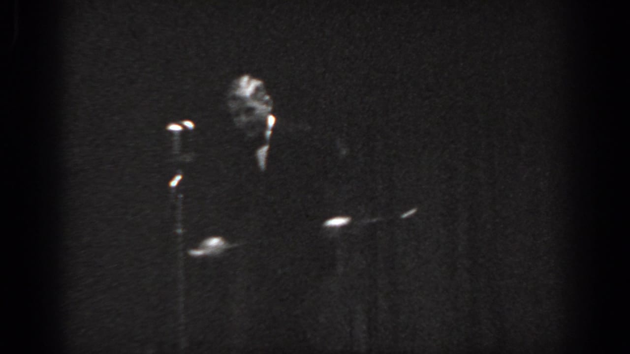 Grainy 8mm black-and-white frame circa 1965, man in dark suit at a standing microphone on an underlit stage, Saint Louis, Missouri.