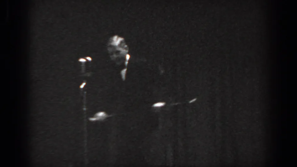 Grainy 8mm black-and-white frame circa 1965, man in dark suit at a standing microphone on an underlit stage, Saint Louis, Missouri.