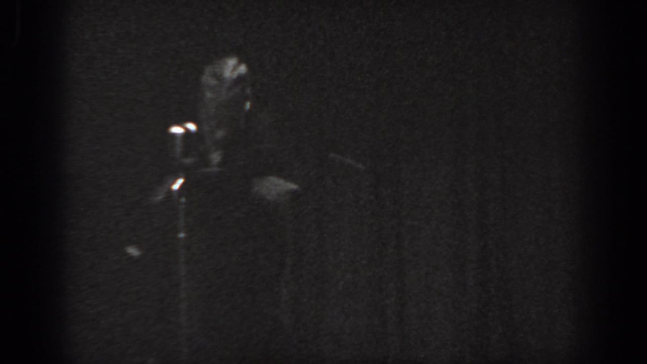 Black-and-white 8mm frame, man at a standing microphone on a darkened stage, curtain backdrop, heavy grain.
