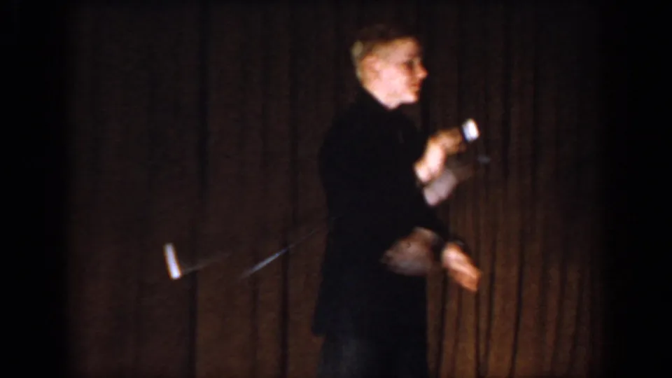 Color 8mm frame circa 1965, male performer in dark clothing gesturing on stage with a brown curtain backdrop, face visible, Saint Louis, Missouri.