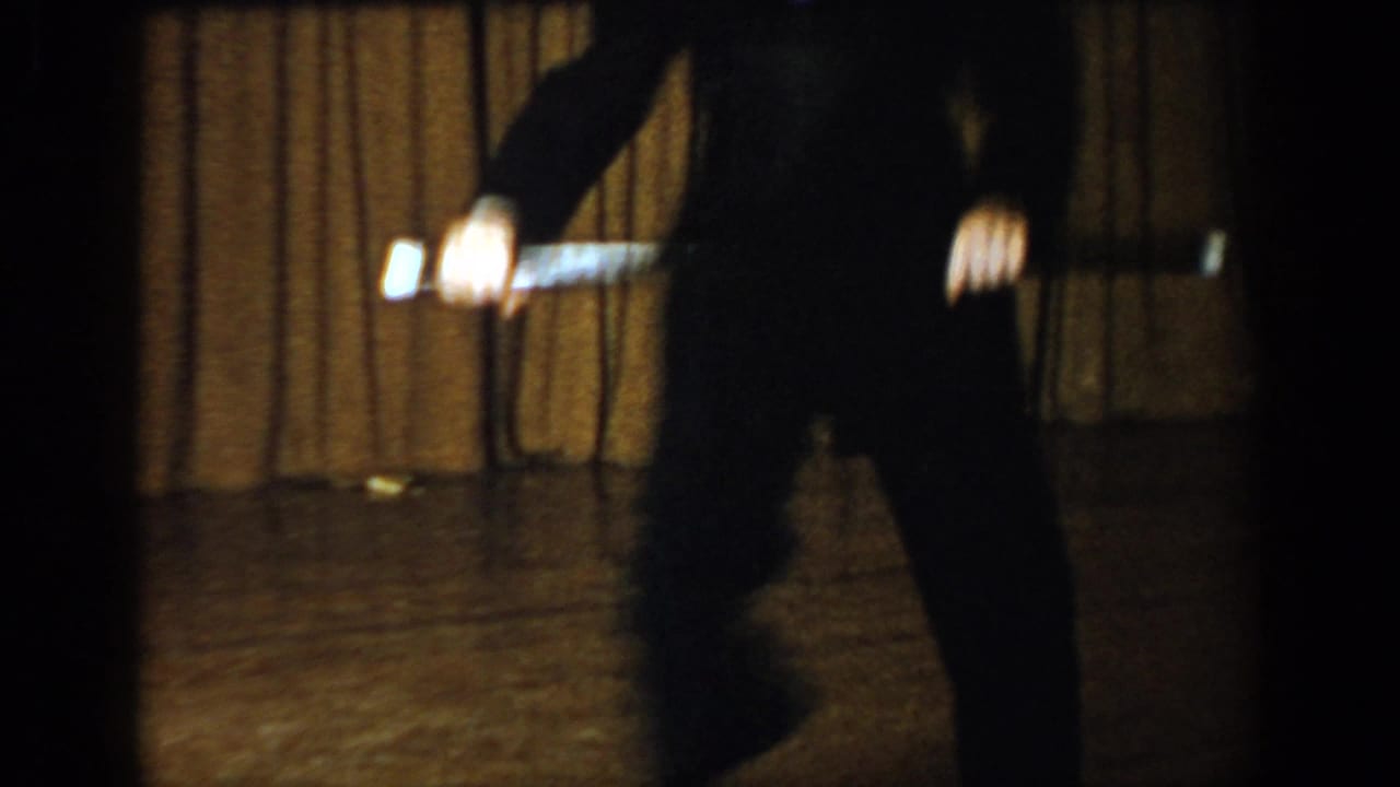 Waist-down view of a performer in dark trousers holding a baton on a dimly lit stage; 8mm footage attributed to Saint Louis, Missouri, c. 1965.