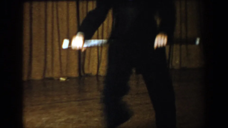 Waist-down view of a performer in dark trousers holding a baton on a dimly lit stage; 8mm footage attributed to Saint Louis, Missouri, c. 1965.