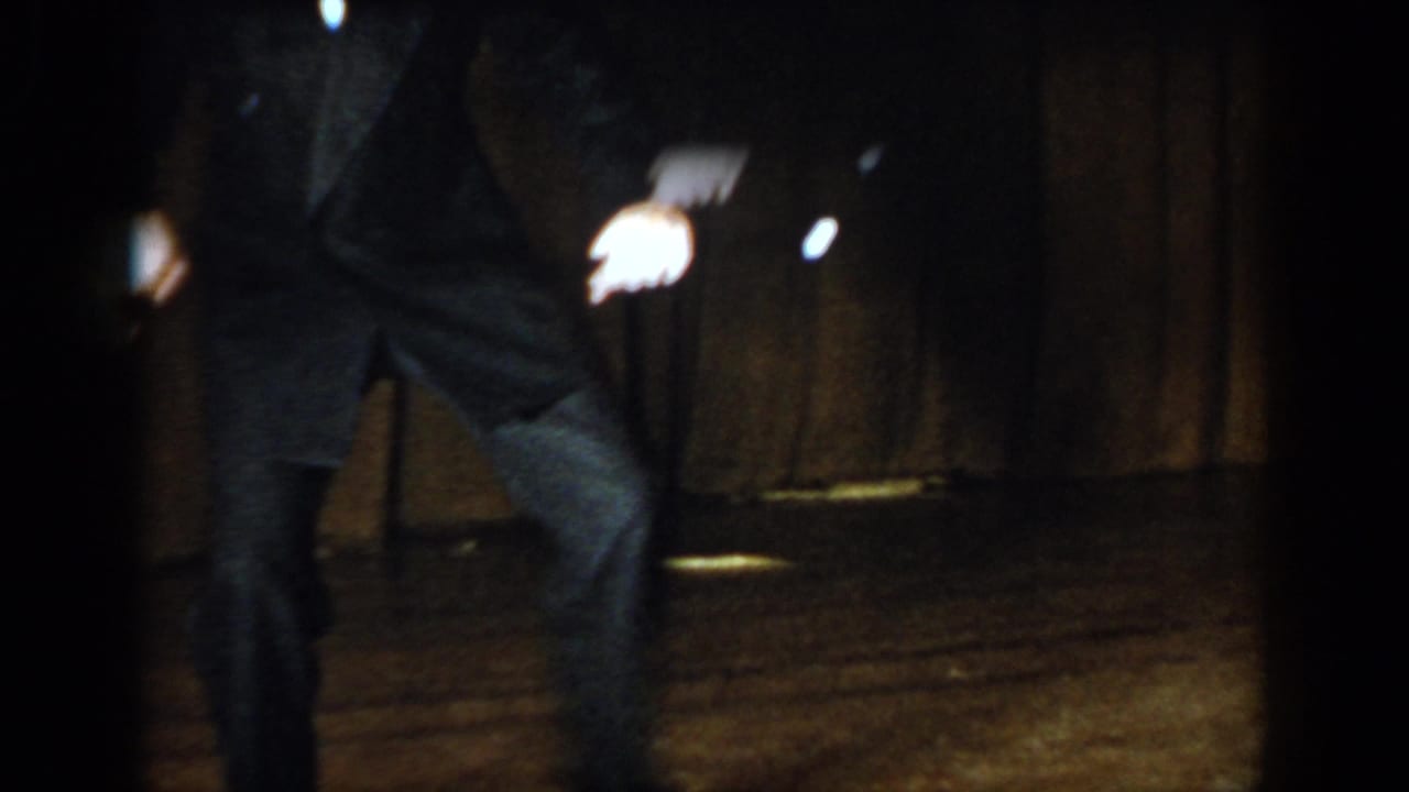 Man in dark suit from waist down with one knee raised mid-dance, holding a baton, brown curtain backdrop on stage.