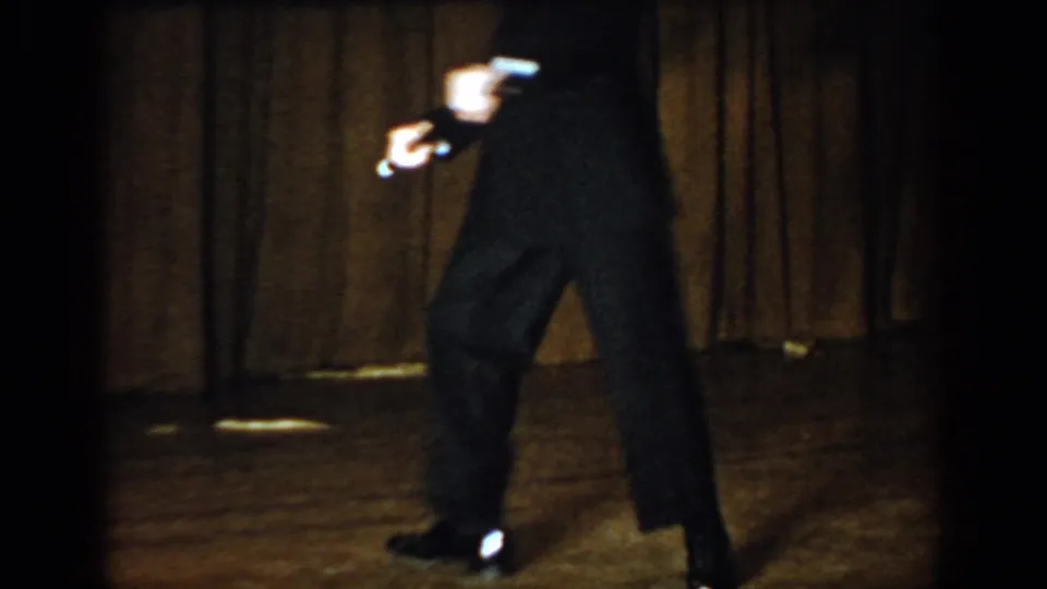 Performer in dark suit leaning into a dance step with baton held low on a wooden stage with draped curtain backdrop; 8mm footage attributed to Saint Louis, Missouri, c. 1965.