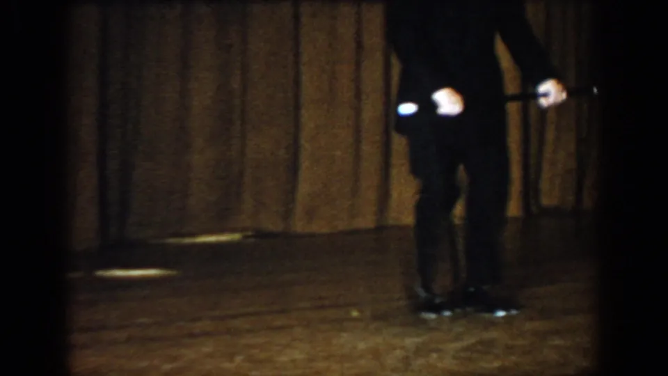Performer in dark trousers standing upright with baton held low at side on wooden stage with curtain backdrop; 8mm footage attributed to Saint Louis, Missouri, c. 1965.