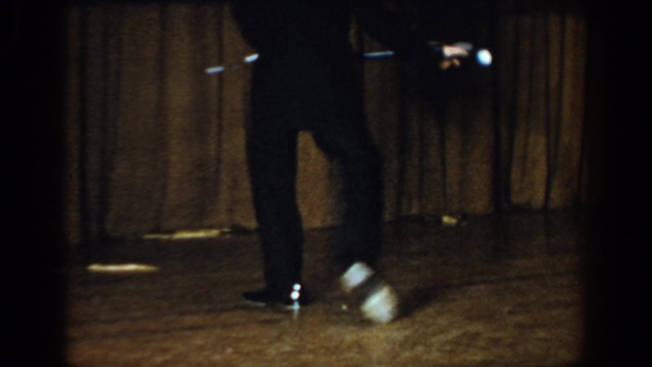 Lower body of a dancer in dark trousers and shoes on a wooden stage, cane held horizontal, feet blurred with motion, brown curtain backdrop.