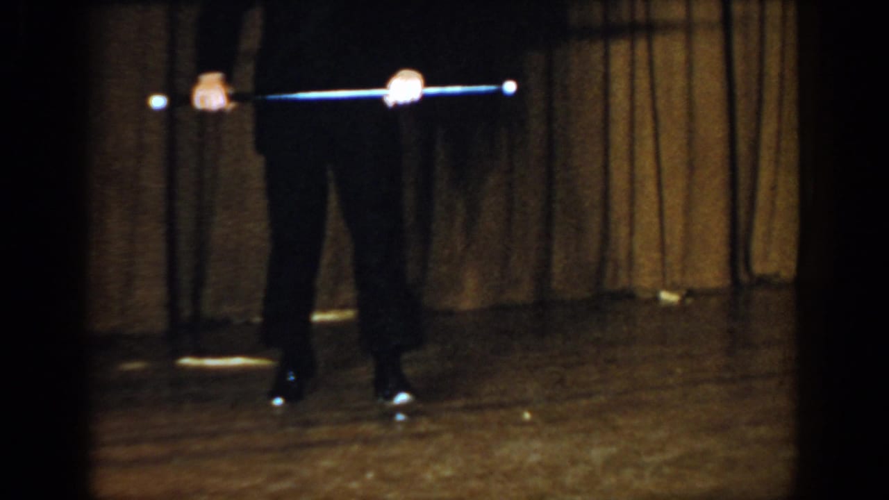 Lower body of a formally dressed figure on a wooden stage, holding a cane horizontally, brown theatre curtain behind.