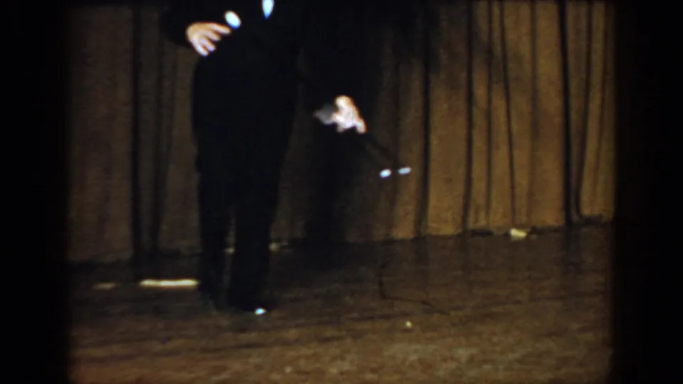 Performer in a black suit crouching on a wooden stage circa 1965, holding a cane angled toward the floor, dim stage lighting and brown curtains behind.