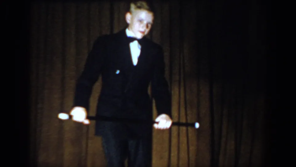 Young performer in a black suit, bow tie, and white shirt on a wooden stage circa 1965, Saint Louis, holding a cane with face clearly visible.