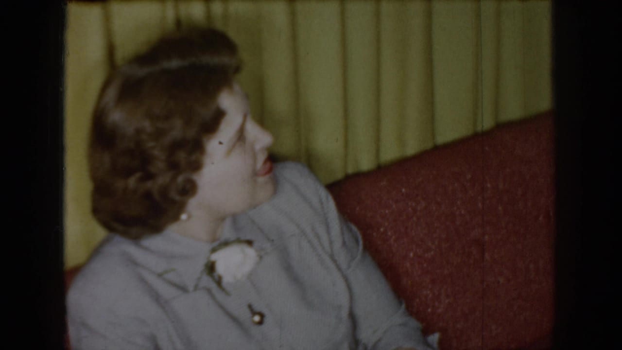 1965 Saint Louis home-movie frame showing a woman in a light jacket looking left while seated on a red couch.
