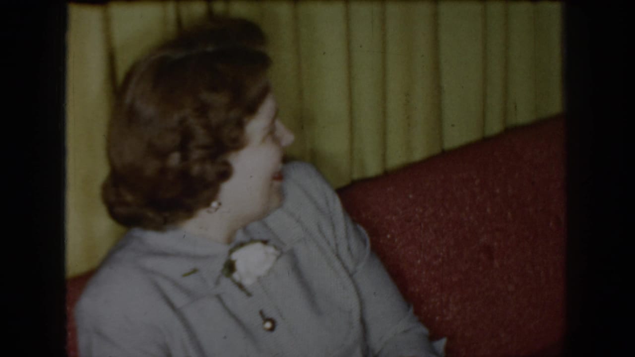 1965 Saint Louis 8mm frame of a woman with curled hair and boutonniere looking to the side from a red couch.