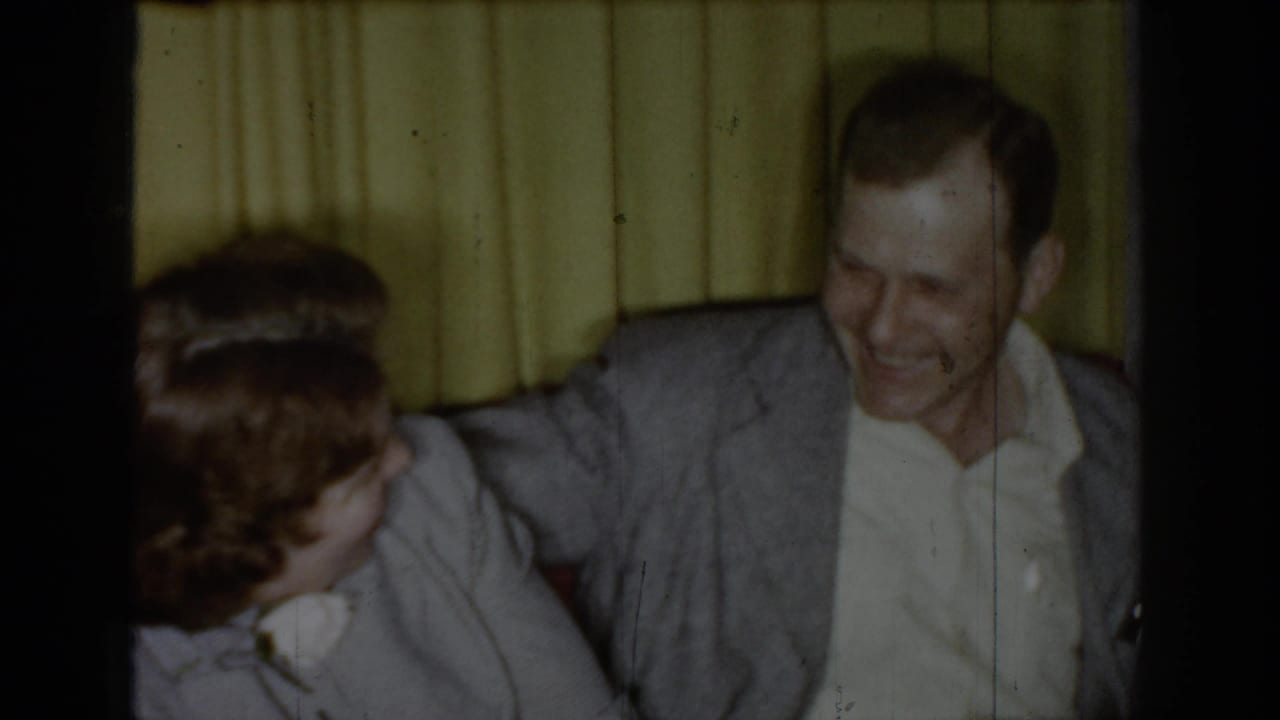 Man in gray sport coat laughing beside a partially visible woman with white corsage in a circa-1965 Saint Louis living room with yellow pleated curtains.