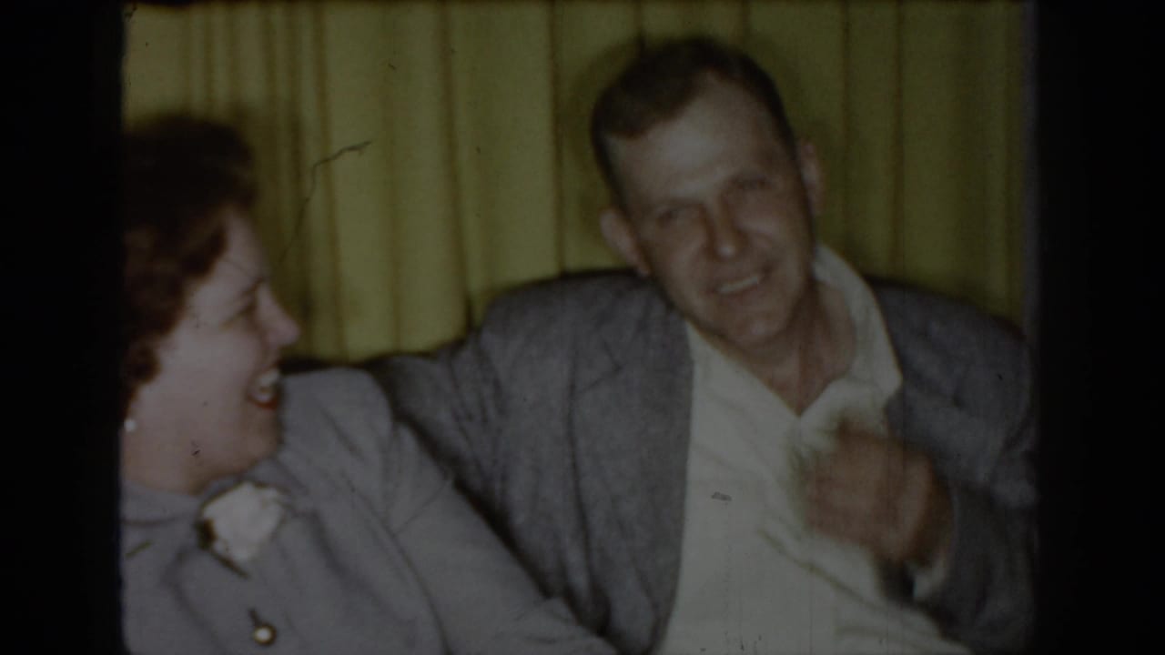 Woman laughing in profile beside smiling man in gray jacket; yellow curtains and hint of red sofa visible.