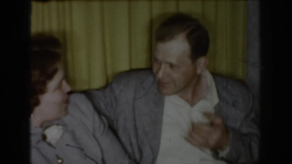 Man in gray sport coat looking down toward a white object while the woman's profile is visible at left; yellow pleated curtains fill the background in a circa-1965 interior.
