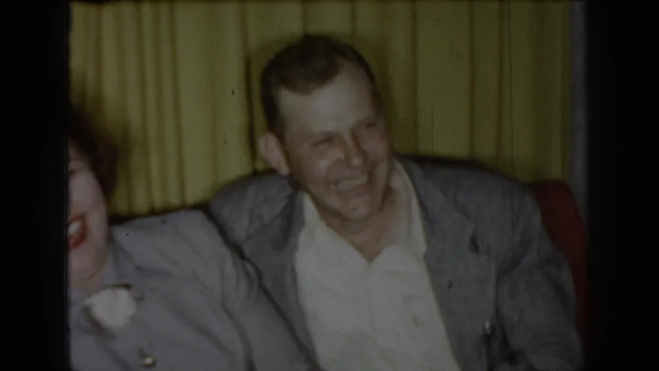 Man in gray jacket laughing openly against a red sofa back and yellow curtains in a circa-1965 Saint Louis living room, woman's head partially visible at left edge.