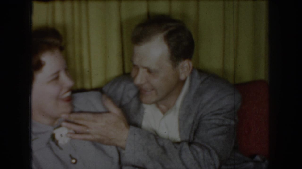 Couple laughing together in a domestic interior, circa 1965 Saint Louis; woman in gray suit with red lipstick, man in gray jacket, yellow drape curtains behind.