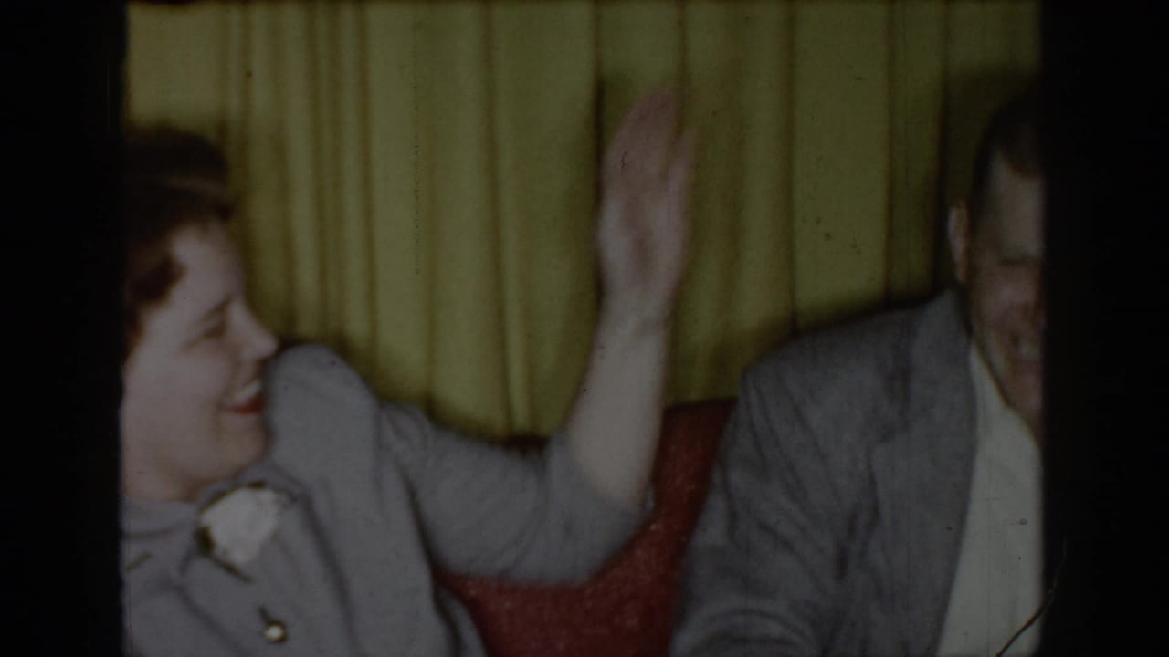 Same couple; woman raises her hand while laughing, man visible at right, yellow curtains and red chair in background.