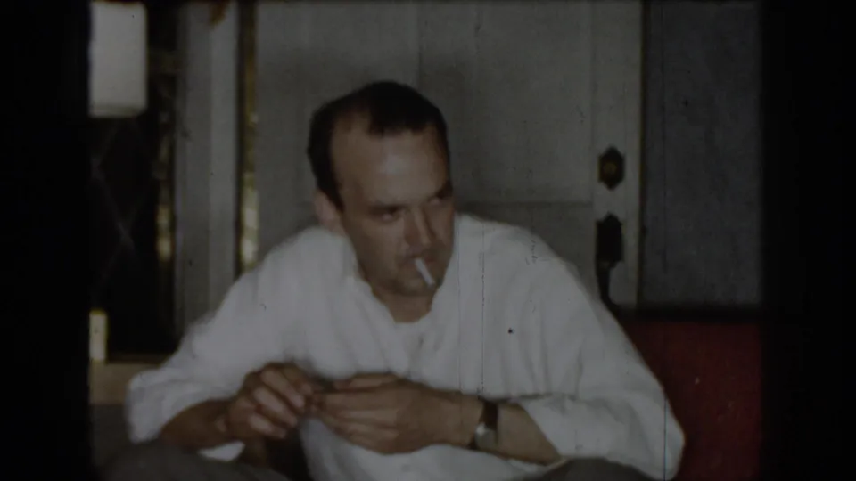 Man in white shirt smoking a cigarette, seated indoors circa 1965; wooden door with metal hardware visible in the background.